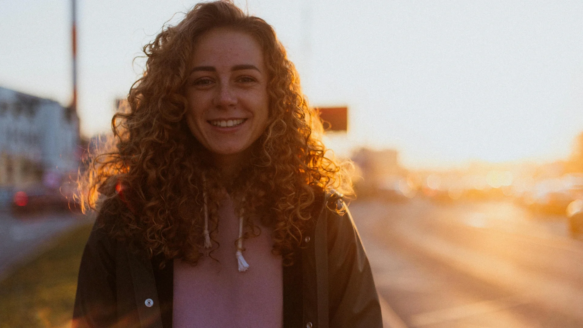 Joyful woman with curly hair smiling in warm sunset light on a city street