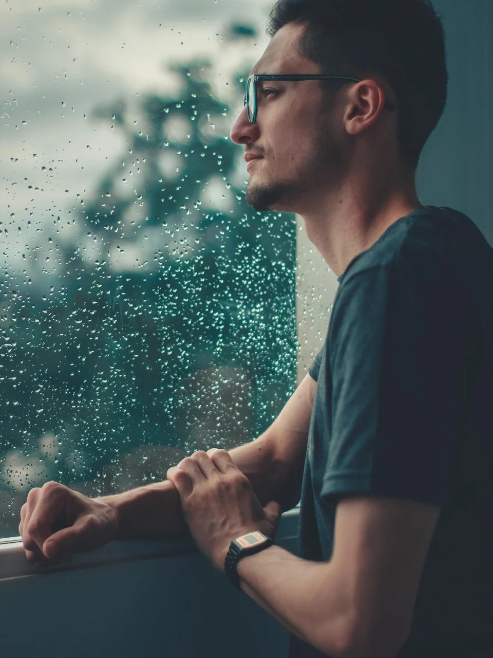 Young man with glasses looking thoughtfully out a rainy window