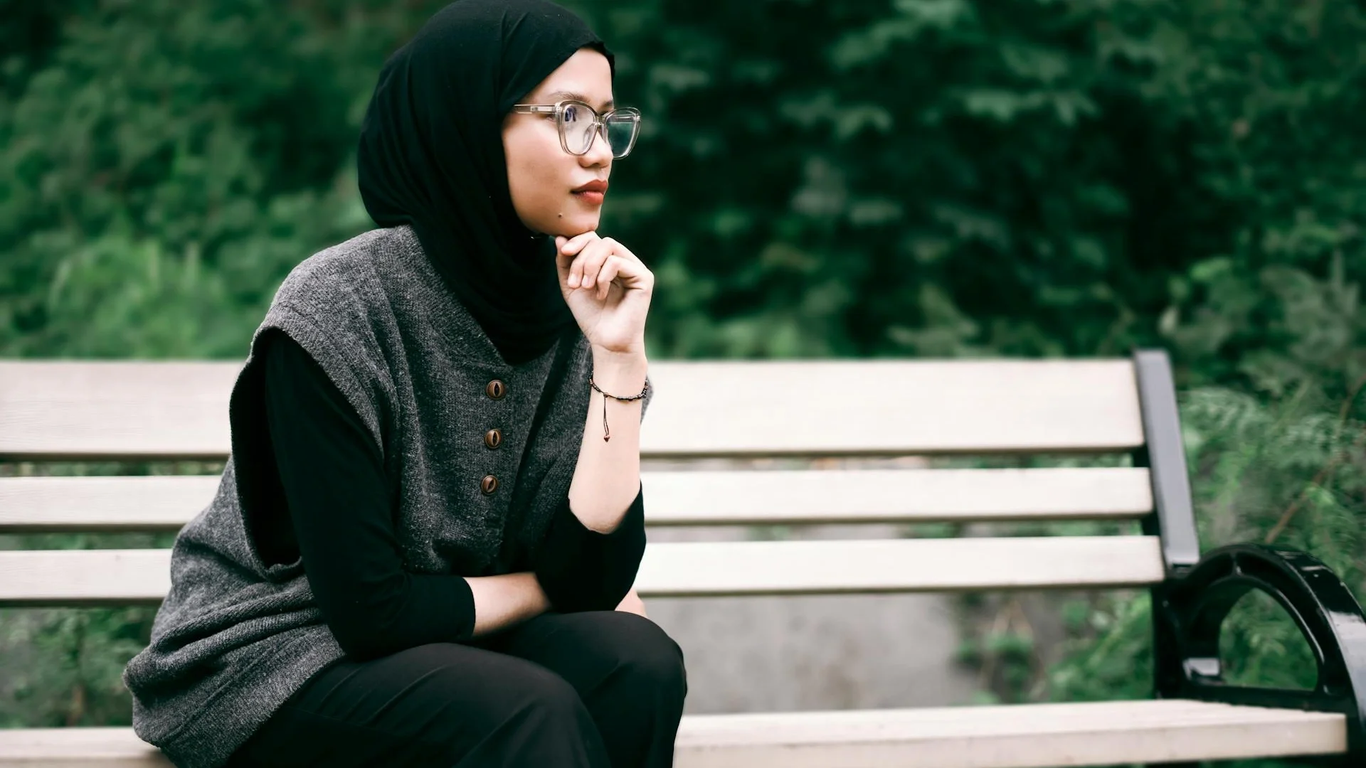 Woman in hijab sitting pensively on a park bench surrounded by greenery