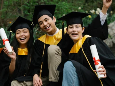 Three diverse graduates joyfully celebrating outdoors with caps, gowns, and diplomas