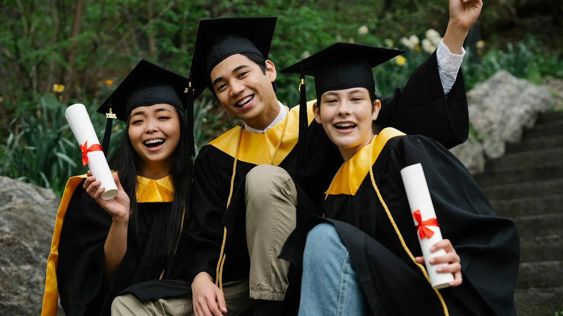 Three diverse graduates joyfully celebrating outdoors with caps, gowns, and diplomas