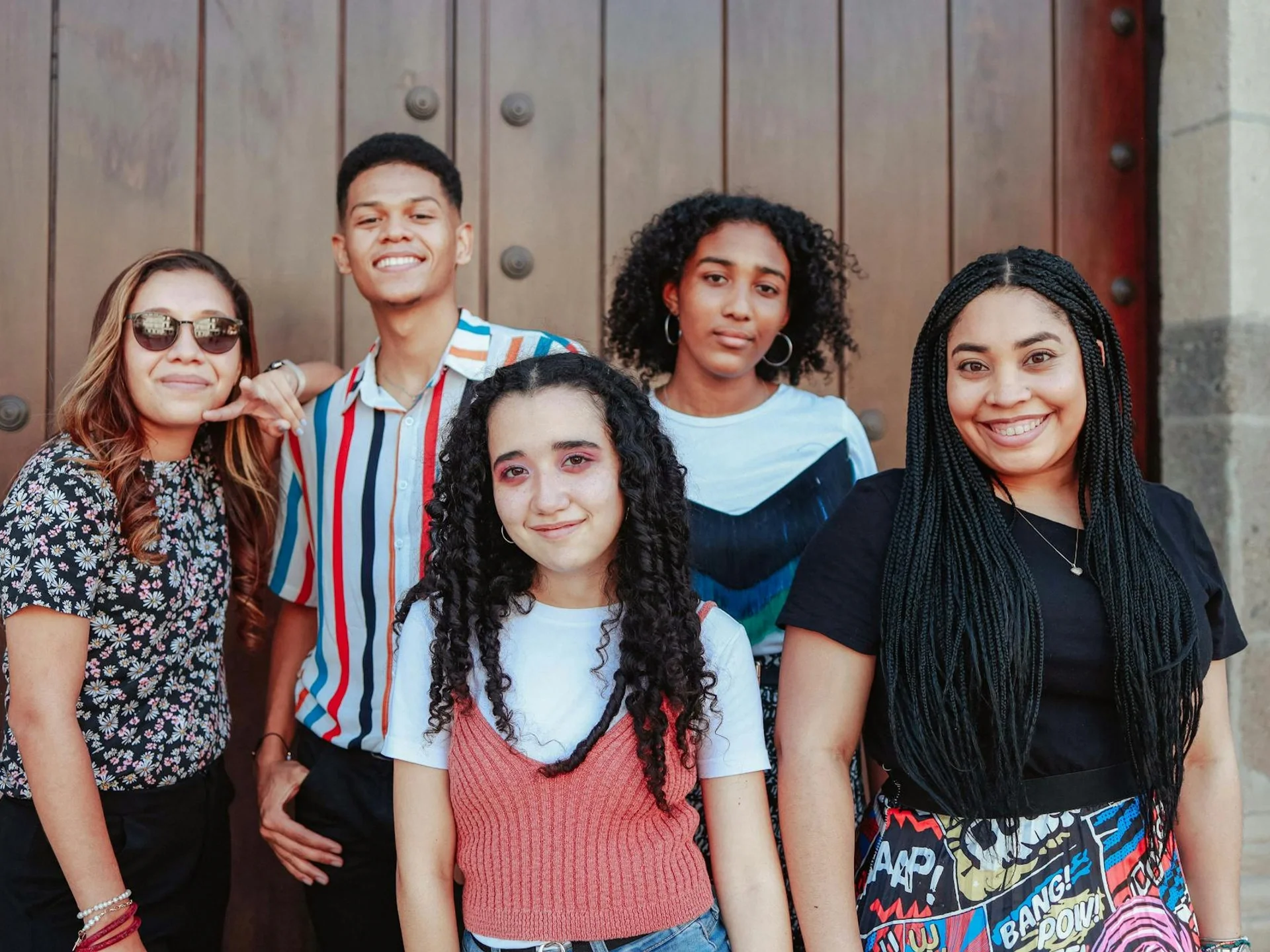 Five diverse teen friends standing together smiling in front of a wooden door